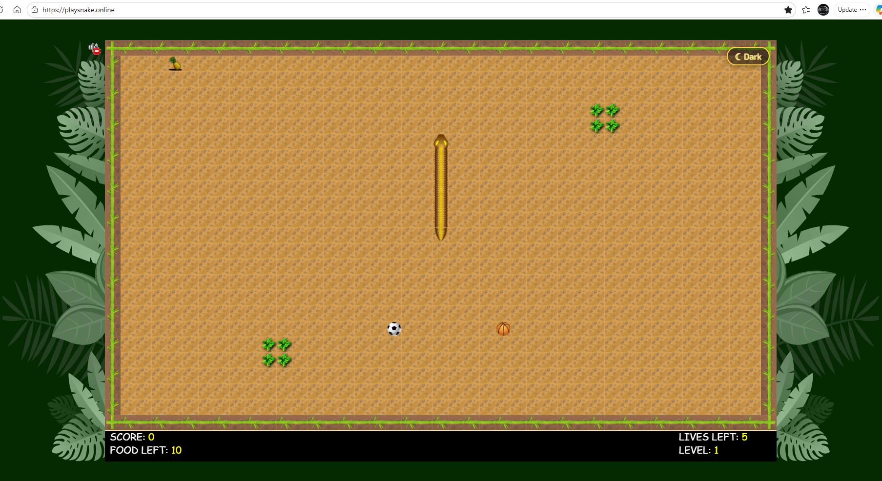 The migrated game in mid-play at playsnake.online: a top-down view of a brown sand arena framed by green bamboo edges and jungle leaves on either side. The yellow snake is moving up the centre. A soccer ball and a basketball sit as food items, a pineapple in the top-left corner, and small green grass clusters as obstacles. A bottom HUD reads SCORE: 0, FOOD LEFT: 10, LIVES LEFT: 5, LEVEL: 1.